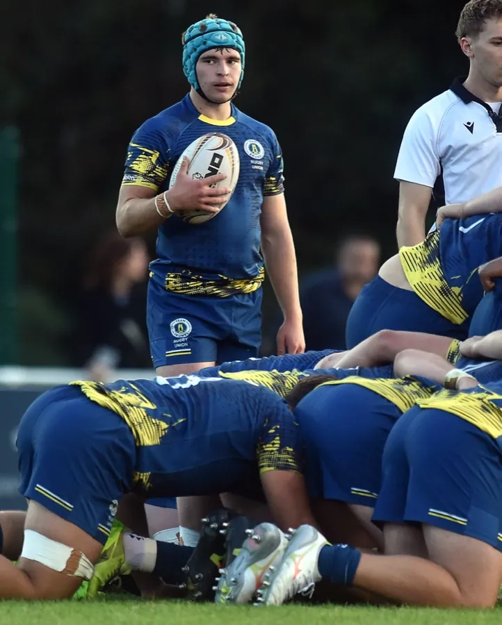 A Brunel scrum-half in a scrum cap stands over the ball at a scrum under floodlights, waiting to feed it in.