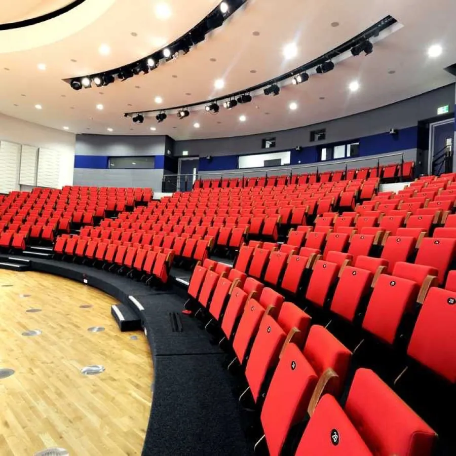 A curved university lecture theatre at Brunel with banked rows of red seats facing a wooden stage floor under stage lighting.