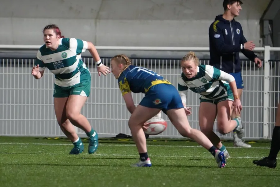 A Brunel women's player in blue and gold bends to gather a loose ball at pace as two opponents in green and white hoops close in.