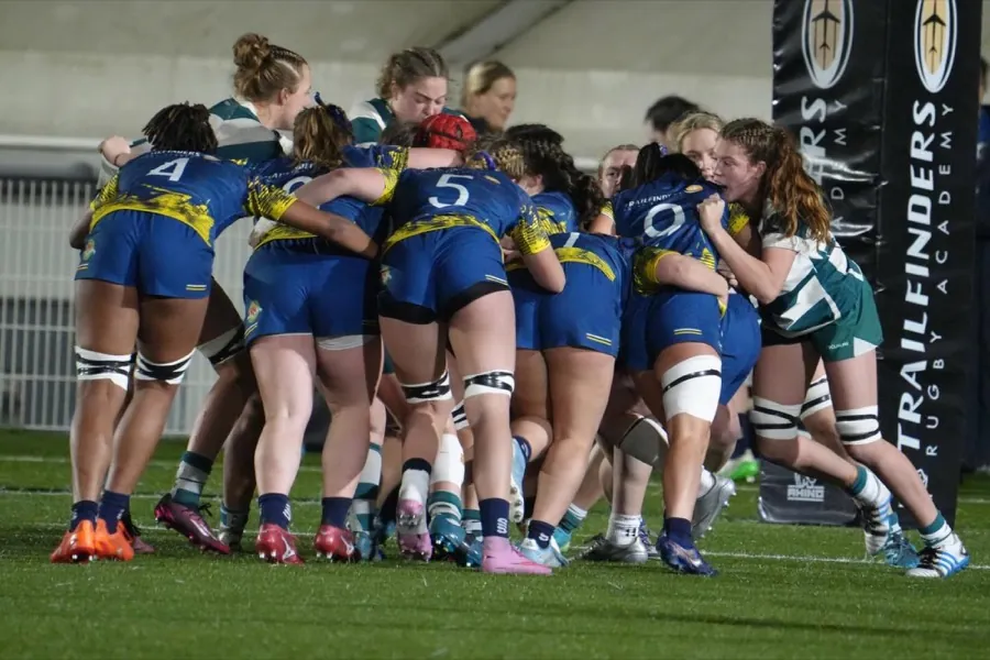 A driving maul under floodlights, Brunel women in blue and gold bound together against opponents in green and white hooped shirts.