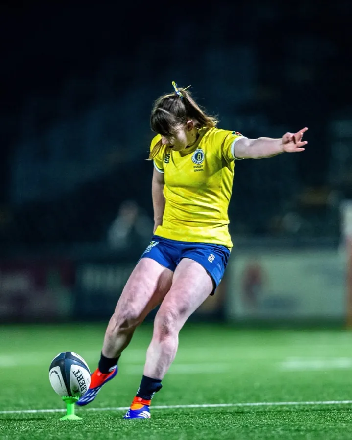 A Brunel women's kicker in the yellow alternate shirt strikes a place kick from a tee on the pitch.