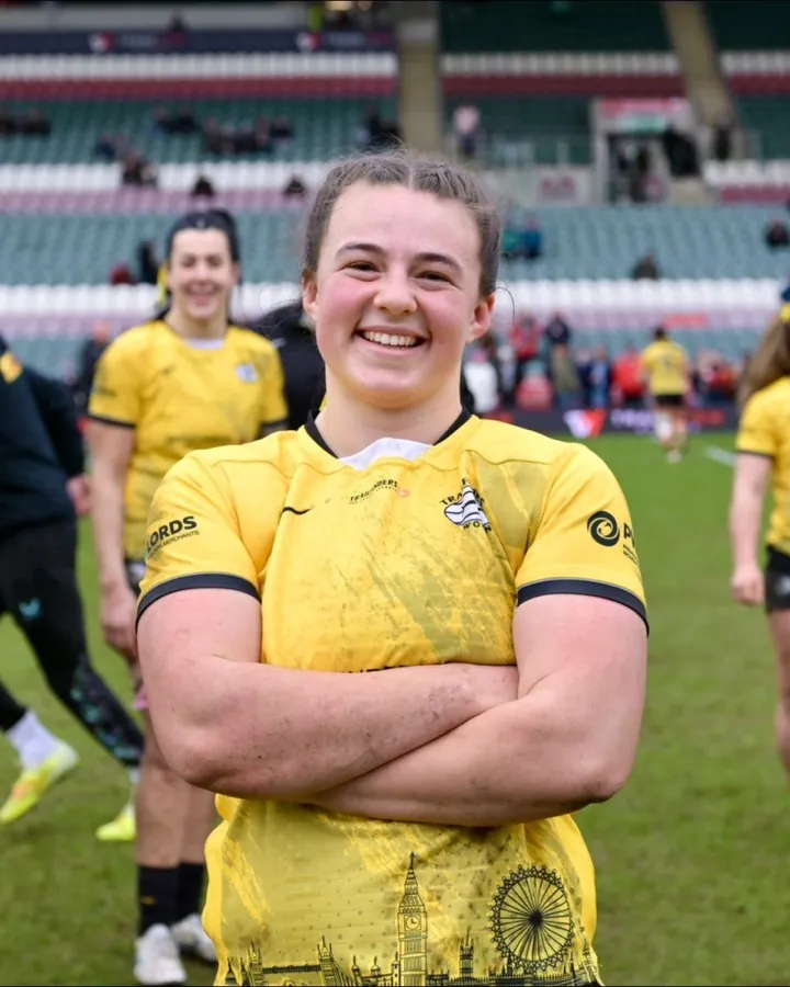 A Brunel women's player in the yellow alternate shirt stands with her arms folded, smiling, with team-mates and a stadium stand behind her.